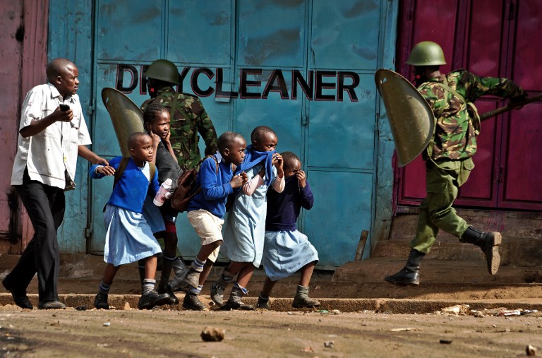 Riot policemen run past school children during a demonstration of opposition supporters protesting for a change of leadership at the electoral commission ahead of a vote due next year, in Kibera slum, Nairobi on May 23, 2016. Local media reported at least one killed in Kisumu in the west of the country, while police in Nairobi and the second city of Mombasa fought running battles with small groups of protesters.There was no immediate police confirmation of the reported death. Police had banned the planned demonstrations and scores of officers in riot gear guarded the building that houses the election commission headquarters in the centre of the capital. / AFP PHOTO / KEVIN MIDIGO